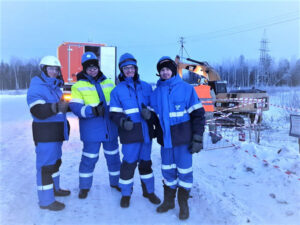 Four workers in winter PPE, safety helmets, and goggles, giving a thumbs-up in snowy conditions