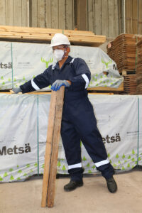 Worker wearing full safety gear, including a hard hat, goggles, gloves, and steel-toe shoes, while handling a wooden plank in a warehouse.